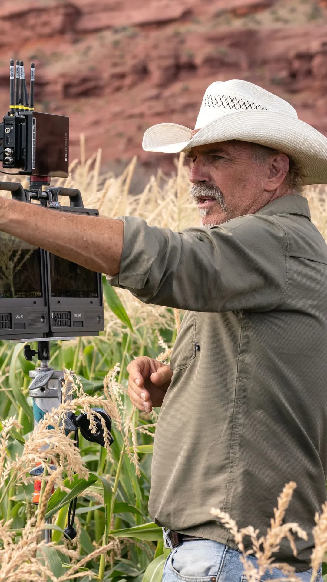 A man in a white cowboy hat and green shirt operates a field drone control panel with screens in a cornfield, red rock cliff behind.