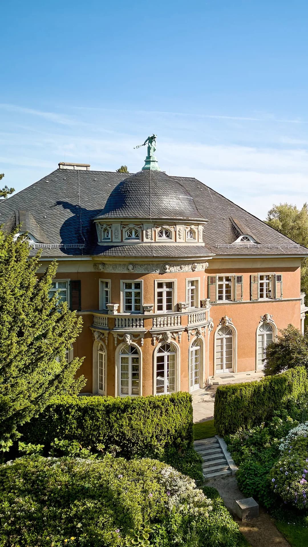 An elegant orange-coloured mansion with a domed tower, ornate windows, a statue on the roof and a lush garden on the riverbank; a bridge can be seen in the distance in the background.