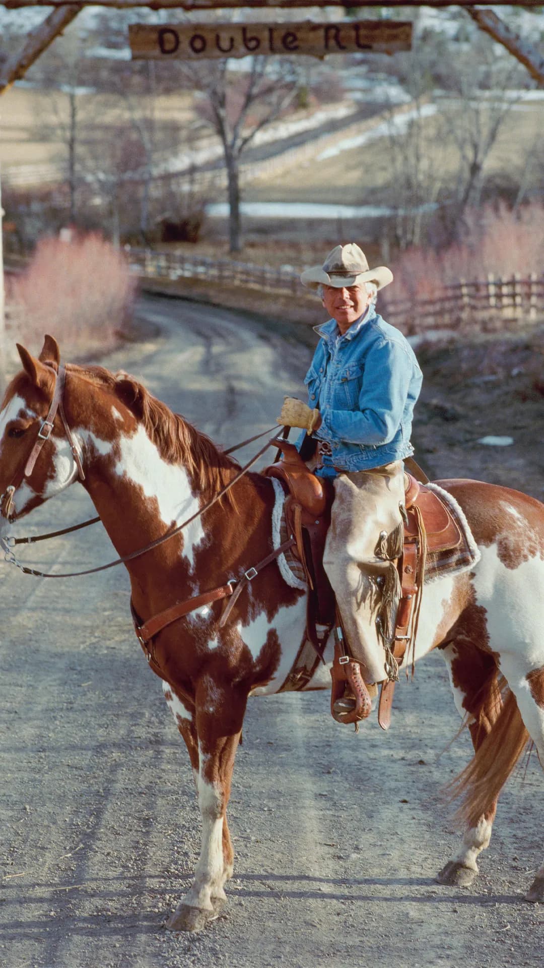 Cowboy in a denim shirt and hat rides a brown-and-white paint horse along a rural dirt road beneath a wooden Double R arch.