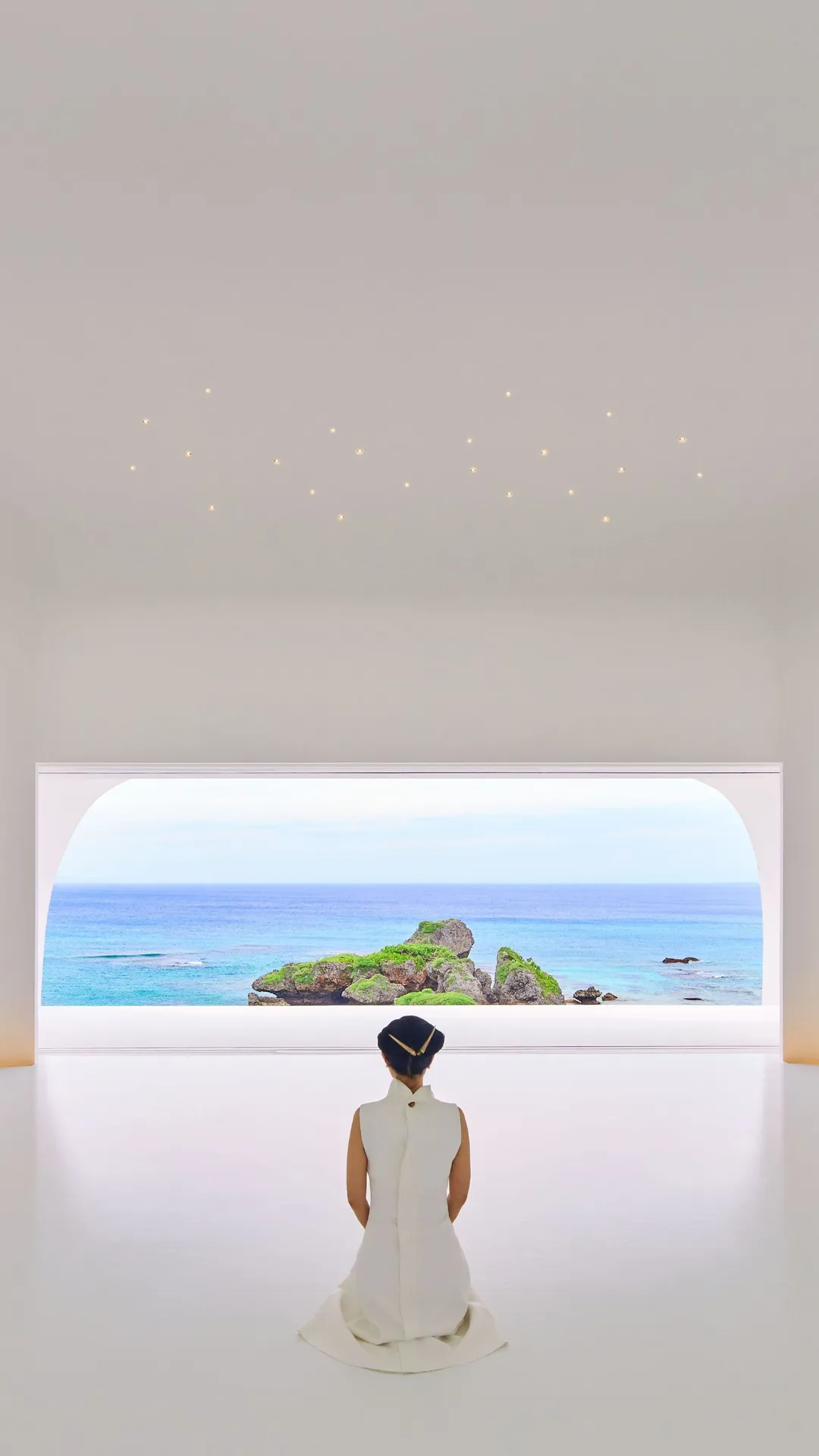 A woman in a white dress kneels in a bright, minimalist room, facing an arched window onto a blue sea and rocky island.