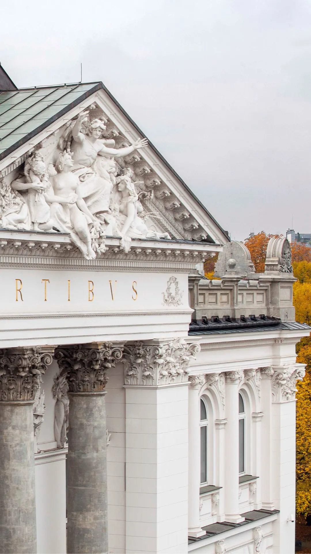 A white neoclassical building with an ornate pediment adorned with mythical figures, tall columns and autumnal trees in the background.