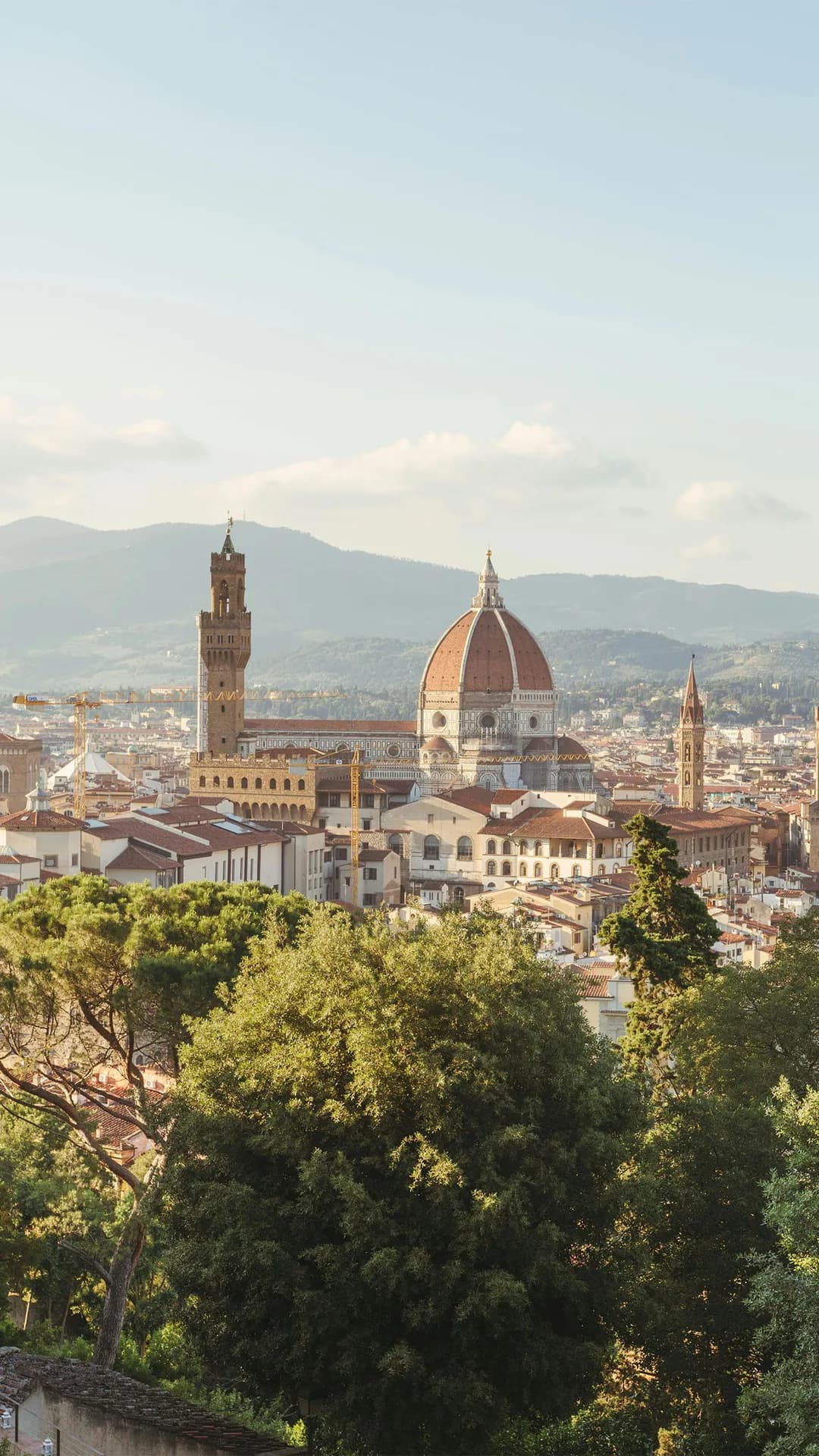 Florence skyline with the Duomo's red dome and historic towers, set against distant hills; foreground filled with green trees.