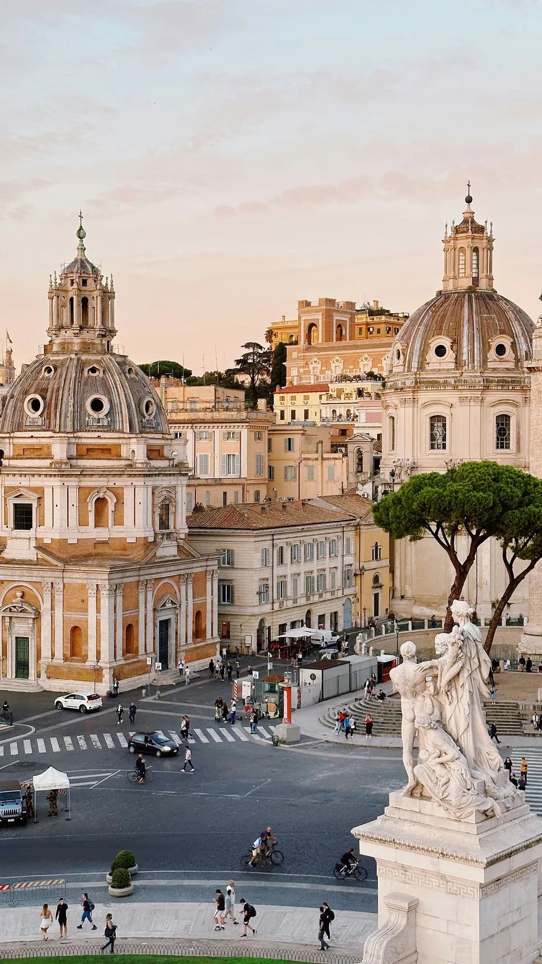 Two domed Baroque churches flank a busy square in Rome, with pastel buildings, trees, and a marble statue in the foreground.
