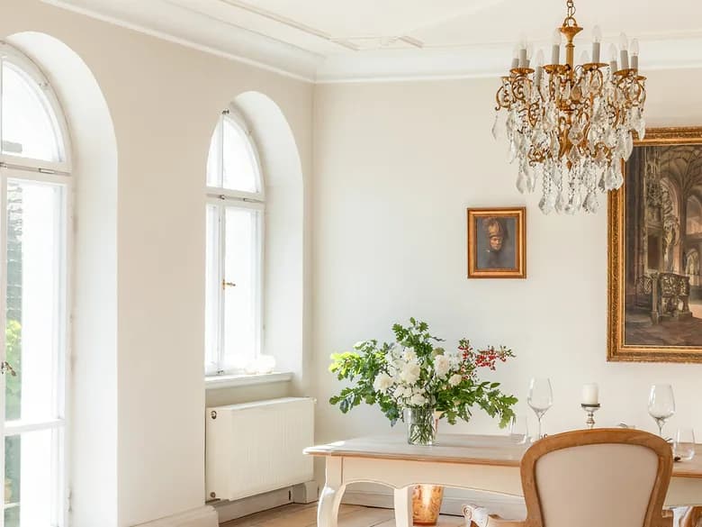 A sun-drenched dining room with decorative ceiling mouldings, a crystal chandelier, arched windows, a wooden table with flowers and a framed painting.