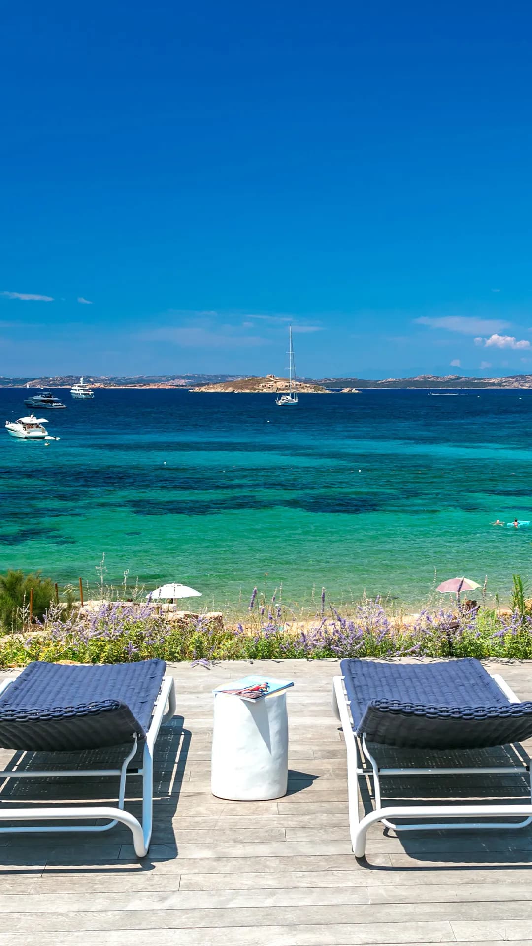 Beachside deck with two lounge chairs, a white side table, purple flowers, turquoise sea, and distant boats under a blue sky.