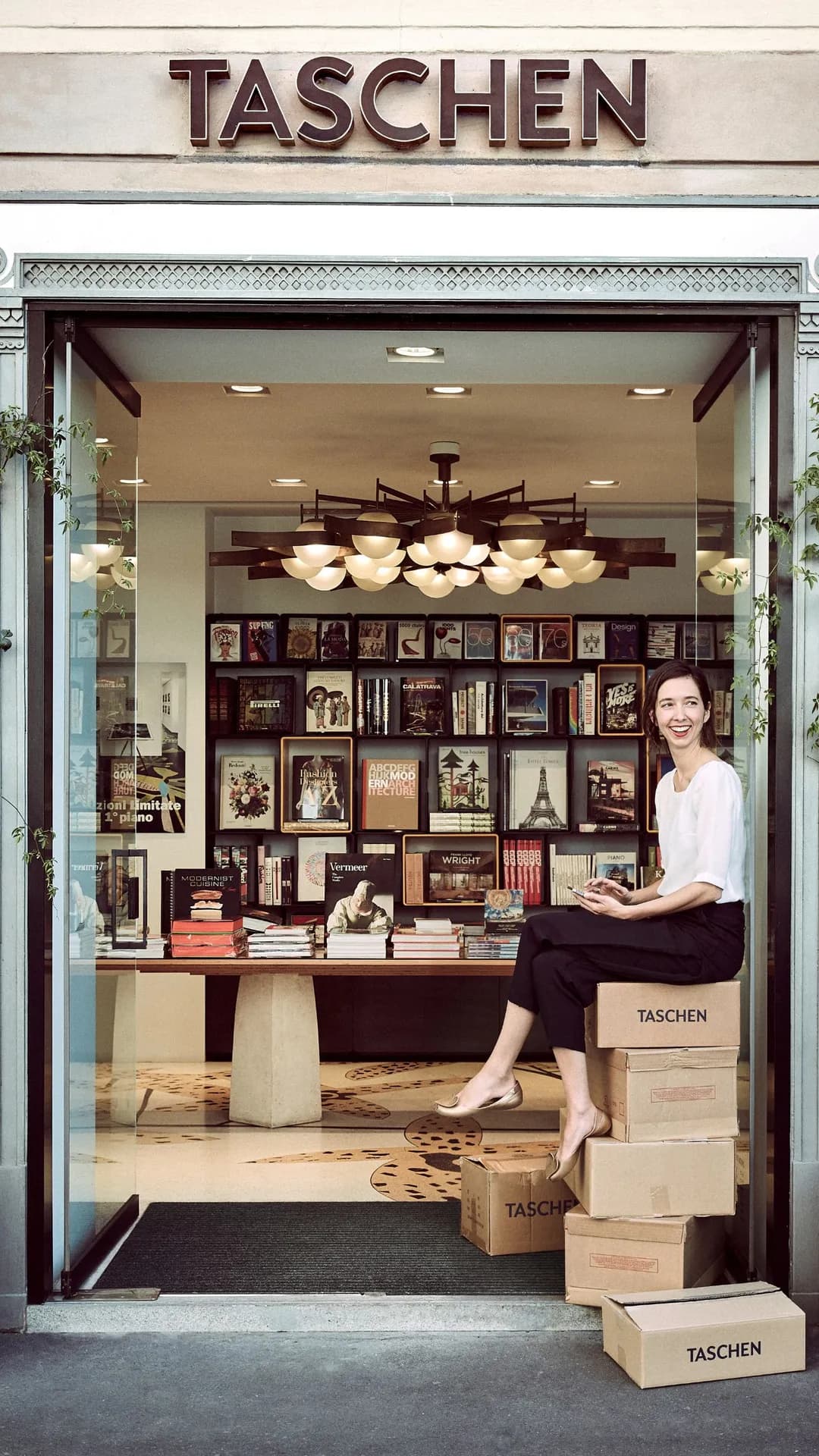 Smiling woman sits on a TASCHEN box at the doorway of a bookstore, with shelves of books and framed art visible inside.
