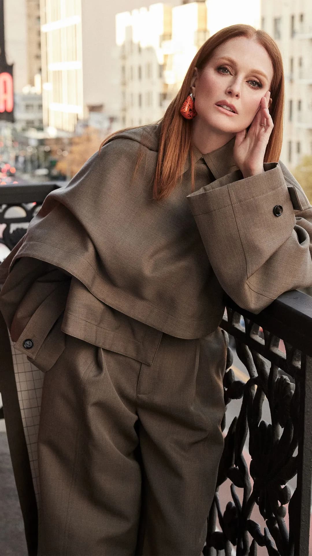 Woman with red hair wearing an oversized taupe coat and matching pants, leaning on a wrought-iron railing against a city street backdrop.