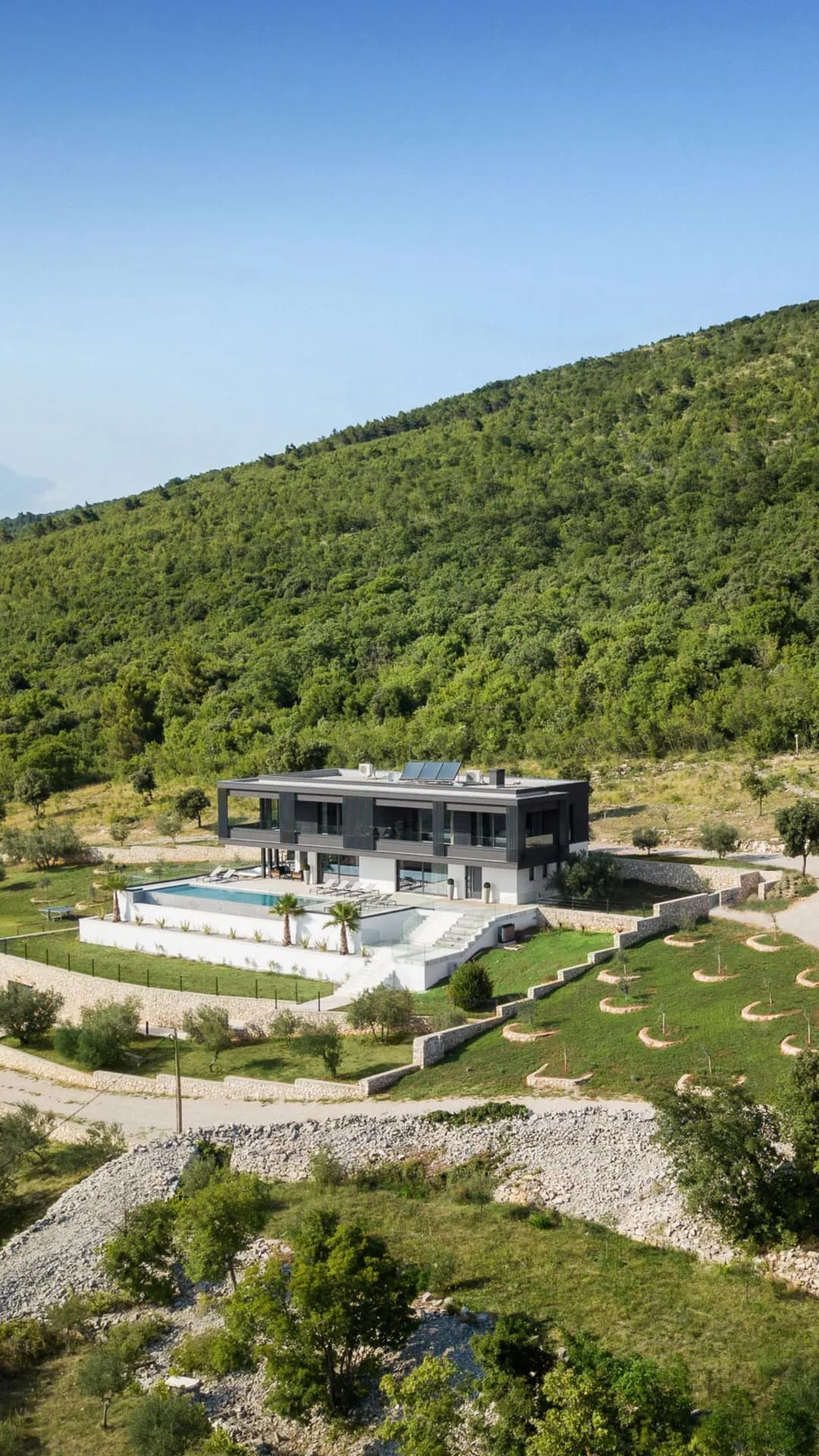 Modern hillside villa with glass walls and a rectangular pool, terraced lawns, and surrounding green forest under a clear blue sky.