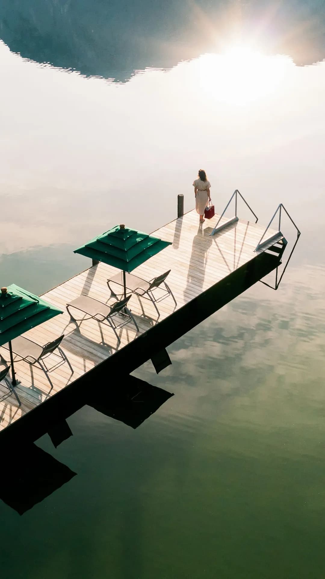 A wooden dock with green canopies and chairs extends into calm water beneath a rocky overhang; a person stands at the end as the sun shines overhead.