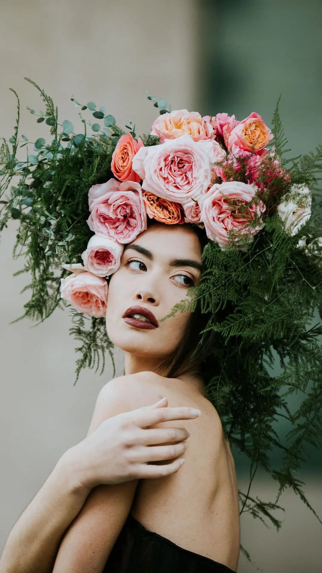 A woman with an extravagant crown of pink roses, ferns, and eucalyptus, gazing to the side with bold lipstick.
