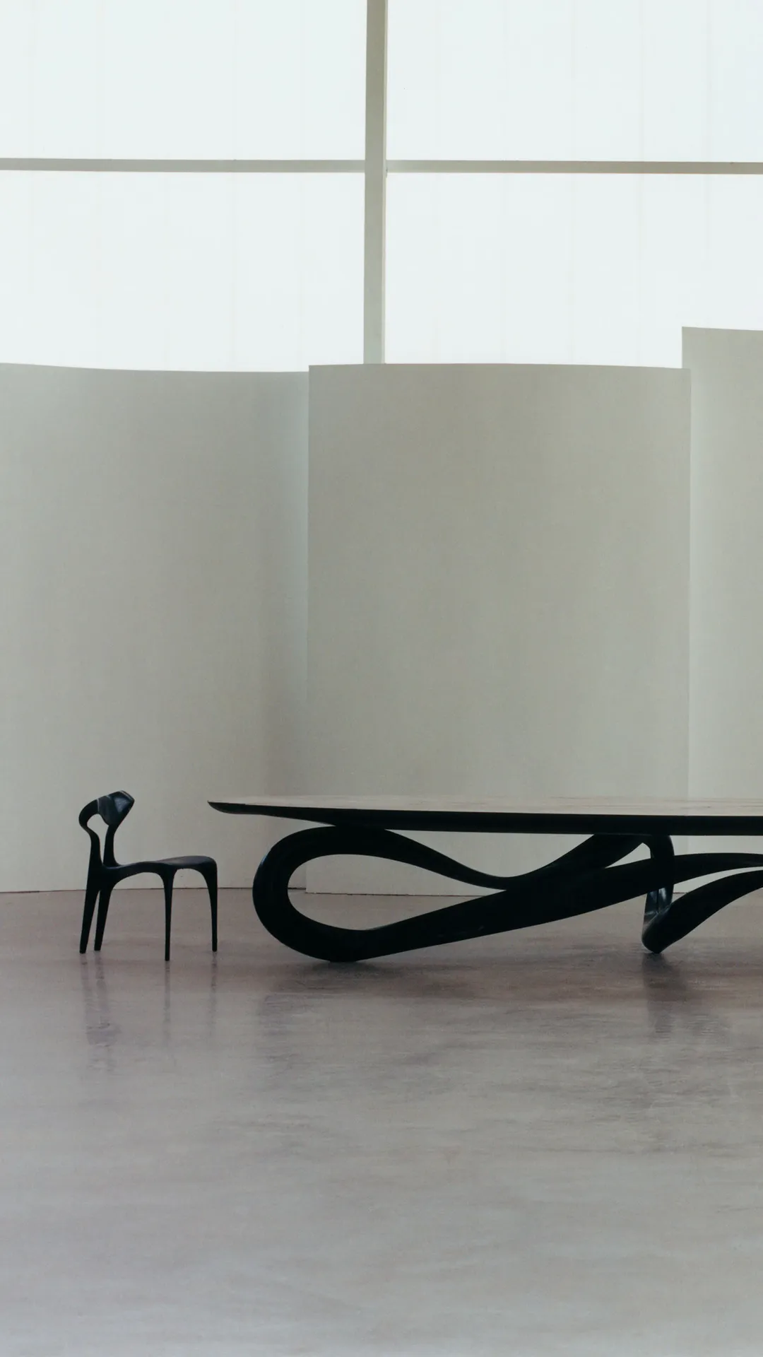 Minimalist gallery with a sculptural black table on looping legs and a small black chair, set against white paneled walls.