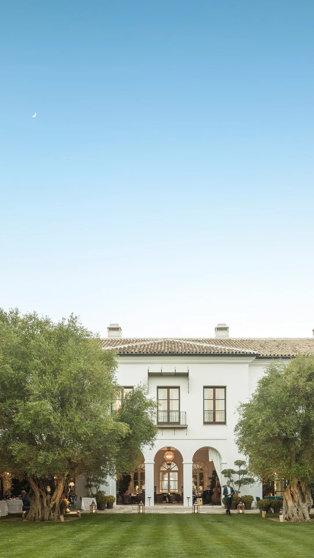 Elegant white villa with arched entrance and terrace dining, surrounded by trees and a green lawn; blue sky with a crescent moon.