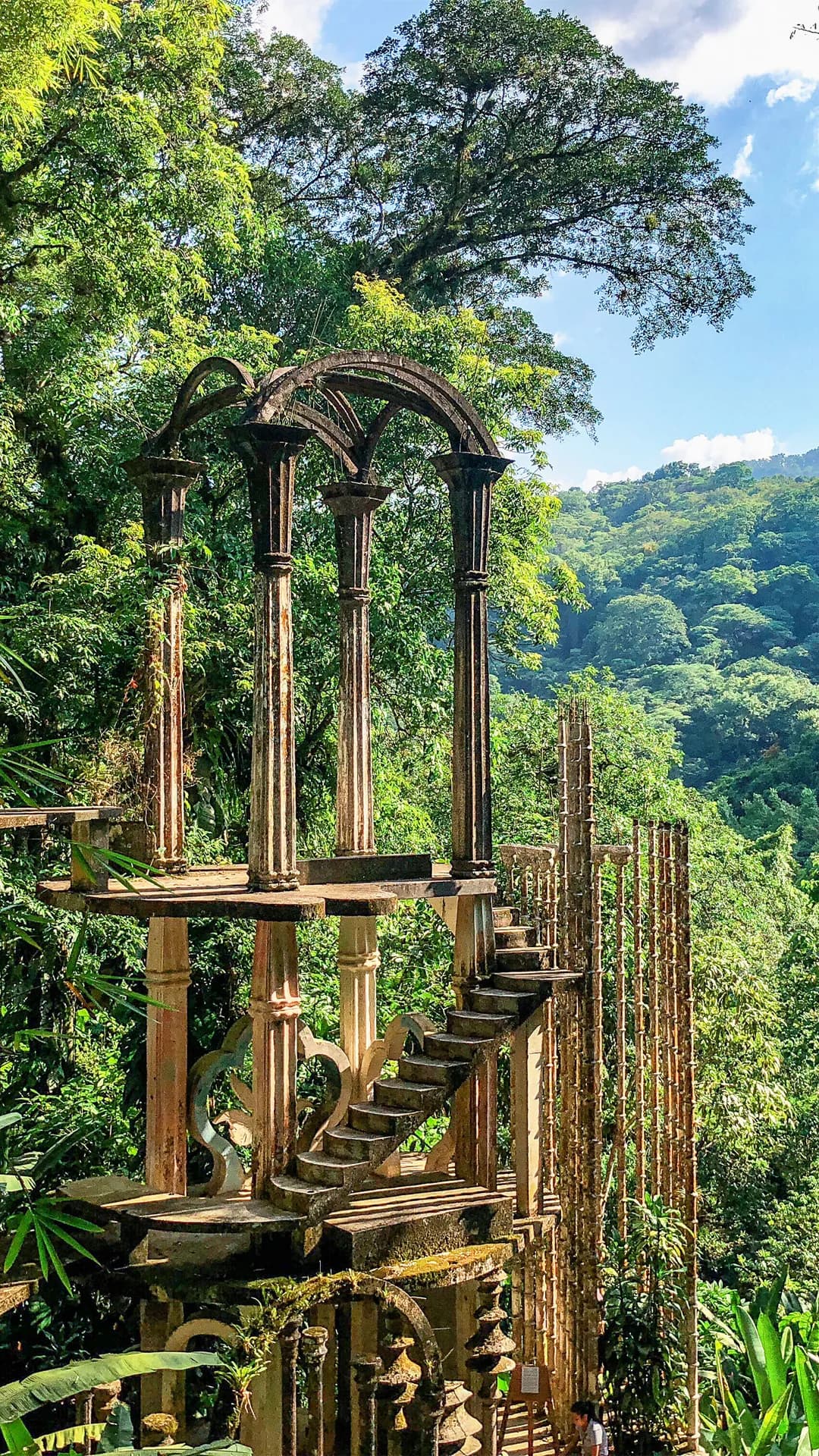 Weathered stone arches and columns frame a stairway through lush tropical forest, with greenery and blue sky beyond.