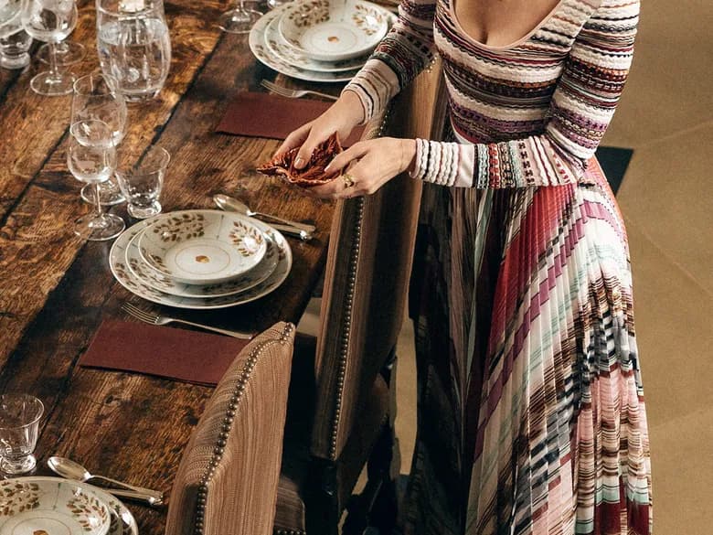A woman in a colourful, striped maxi dress is standing smiling next to a rustic dining table, holding a red object in her hand; there are plates and glasses on the table.