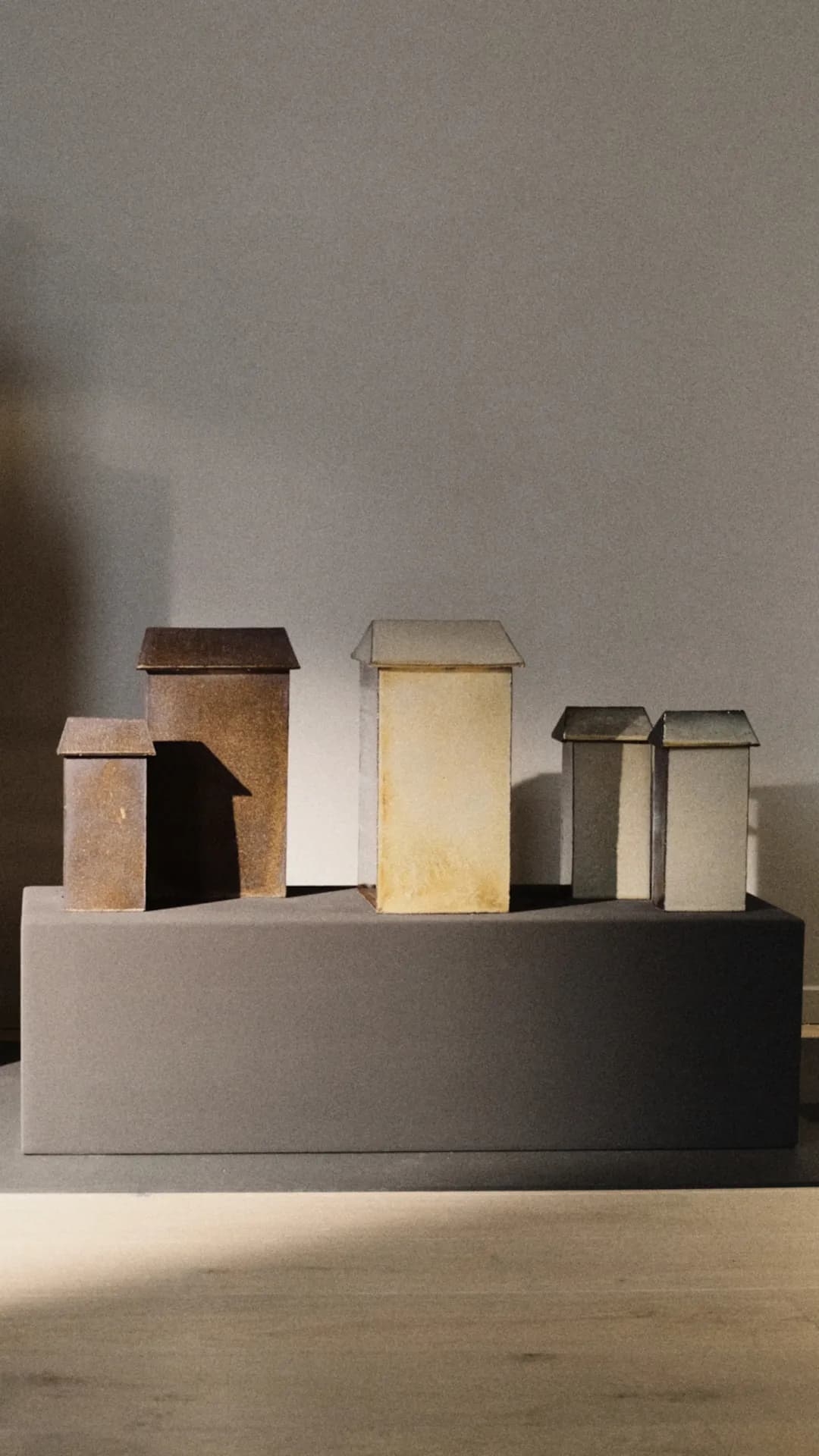 Six small cardboard houses with slanted roofs in brown, tan, and gray hues sit on a gray pedestal.