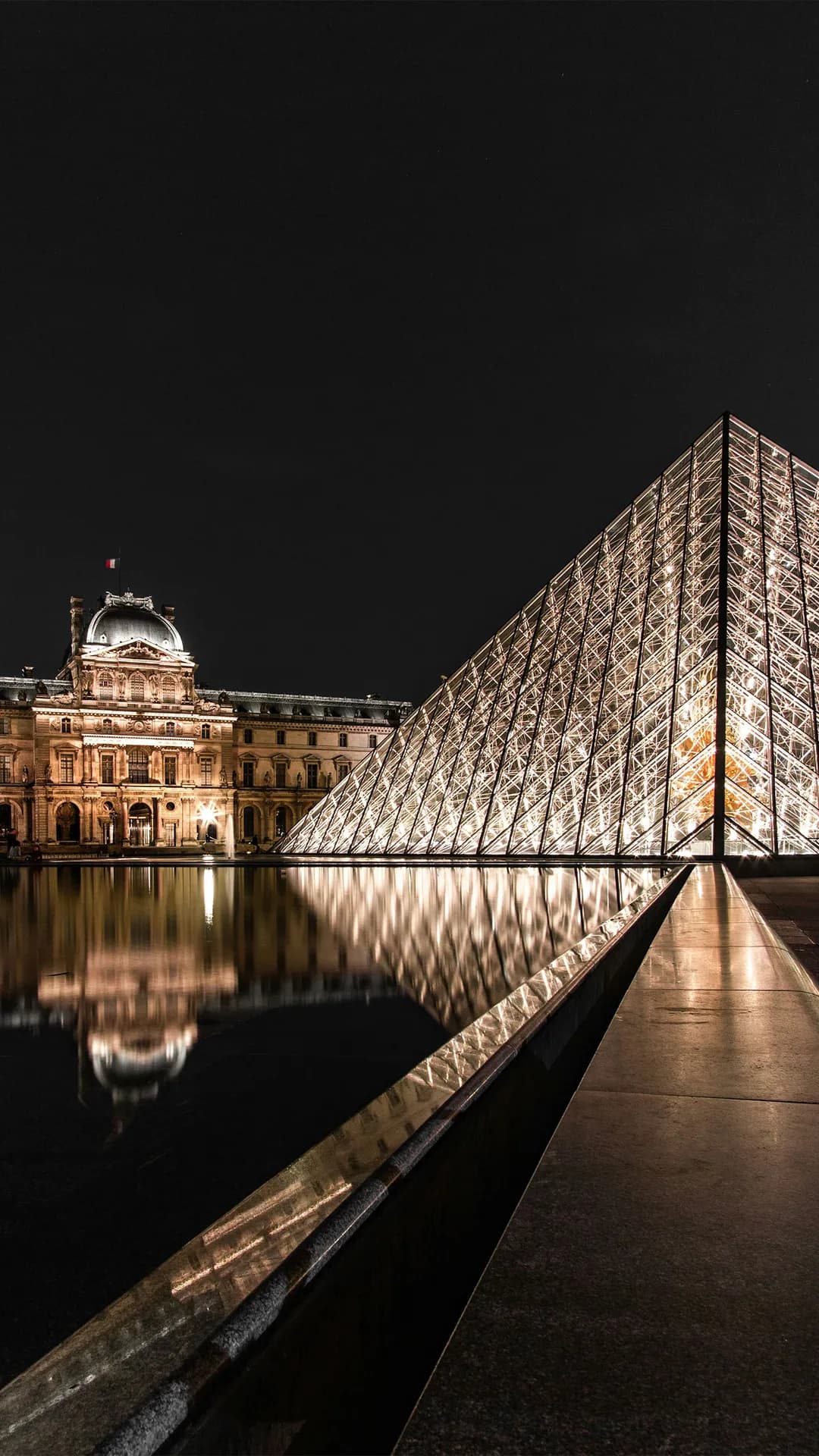 Night view of the illuminated Louvre Pyramid beside the historic Louvre Palace, with glass panels glowing and reflecting in the water.