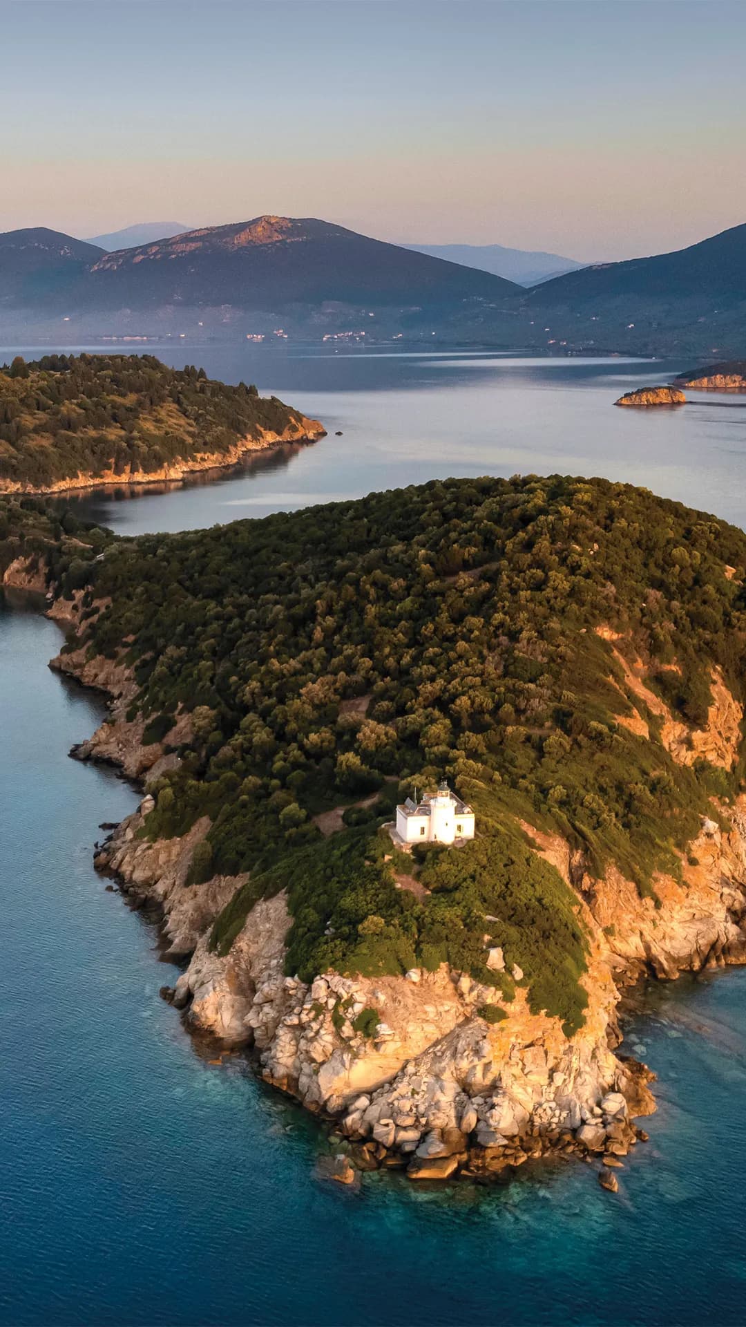 Small white house perched on a rocky, green island surrounded by calm blue water, with distant mountains and a pastel sunset.