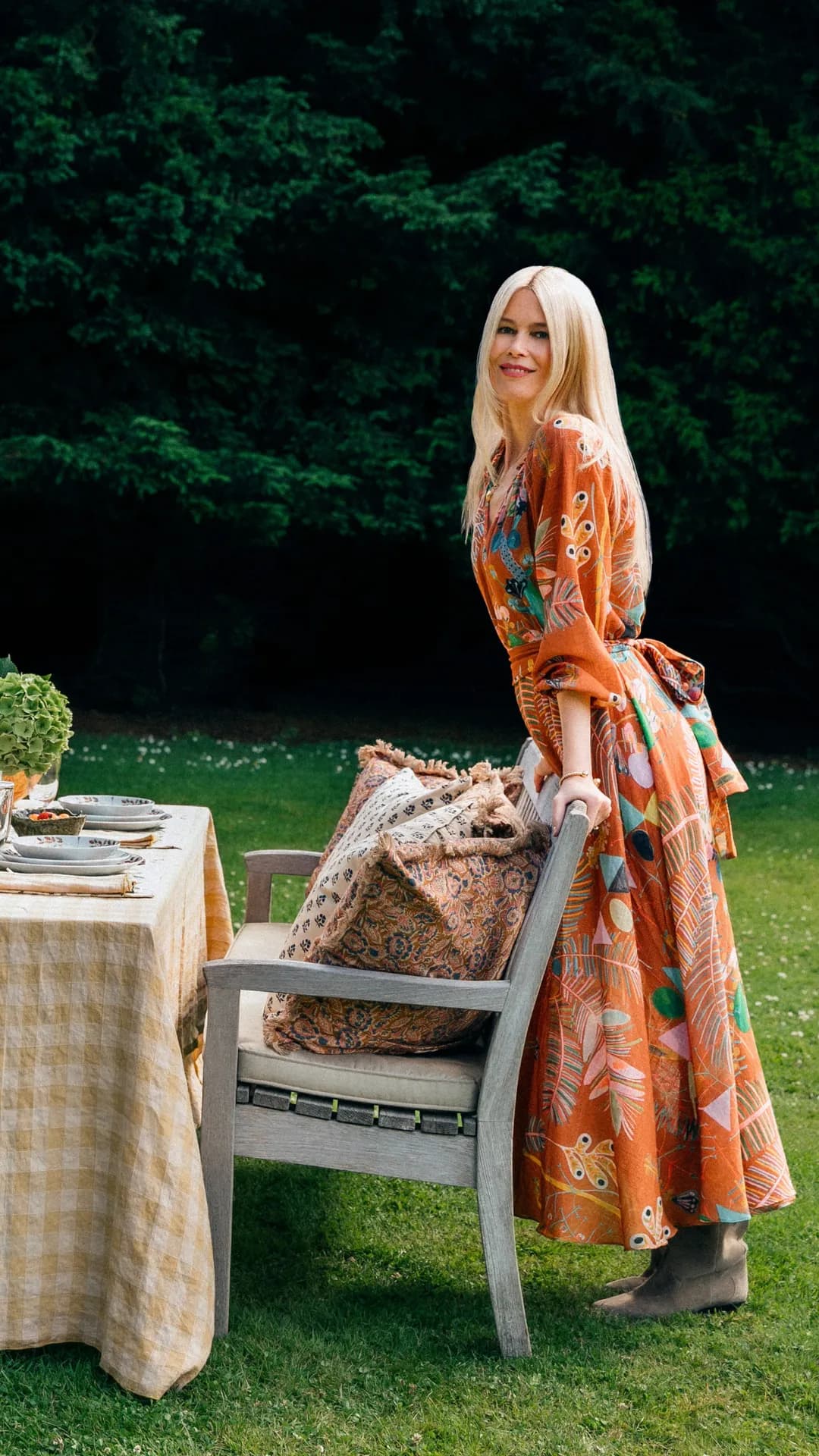 Blonde woman in an orange patterned dress leans on a chair beside an outdoor dining table set on the lawn with cushions and tableware.