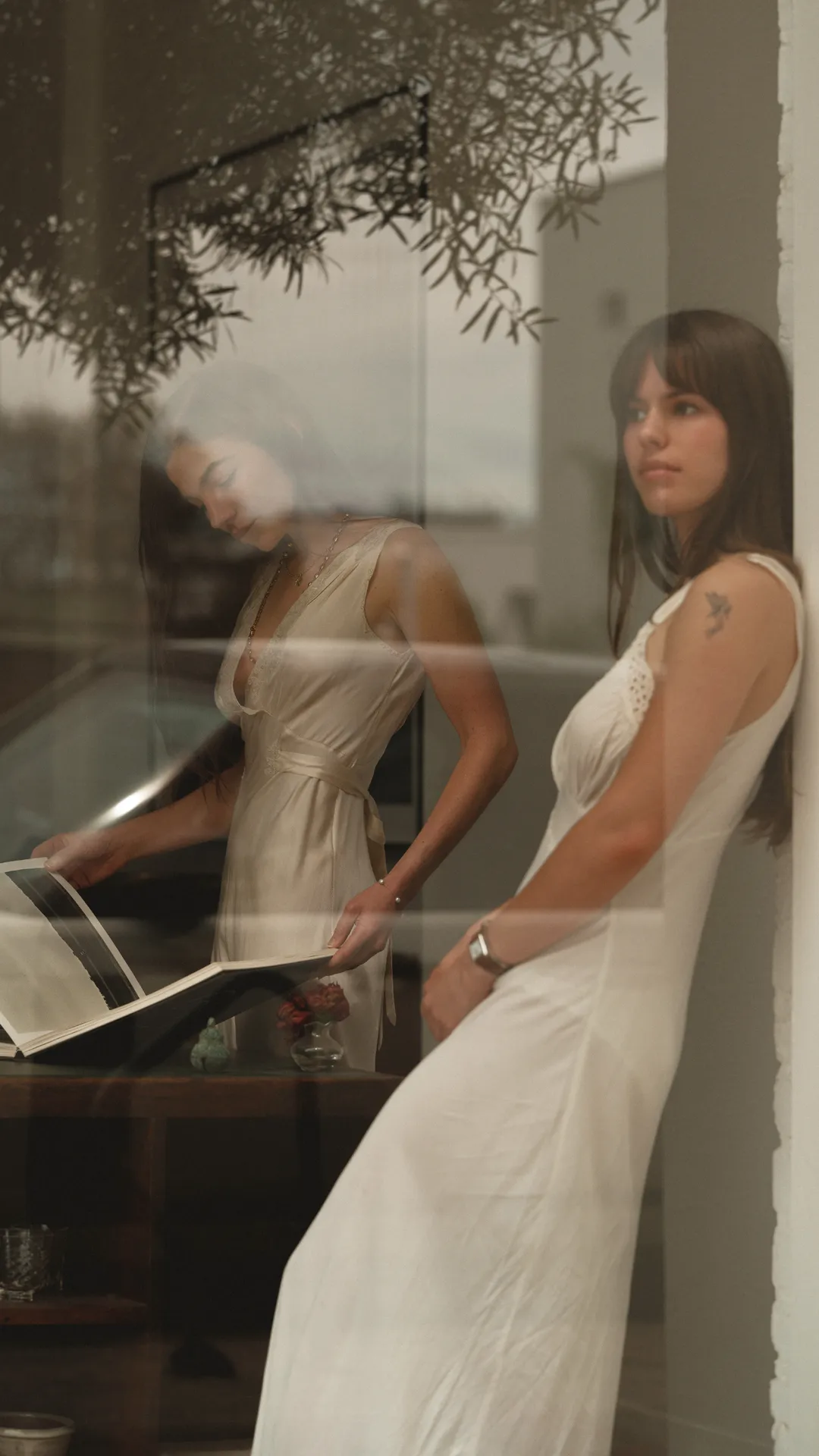 Two women in white dresses by a window, one reading a photo album on a table, the other leaning against the wall, with a glass reflection.