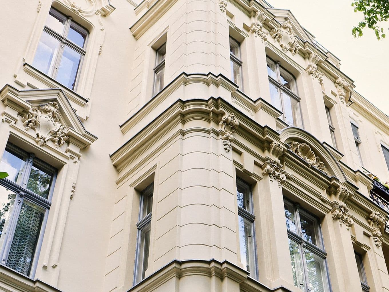 Ein hell cremefarbenes, historisches Wohngebäude mit reich verzierten Fassadendetails, darunter dekorative Fensterrahmen, skulpturale Ornamente und gestufte Gesimse. Hohe, schmale Fenster spiegeln den Himmel und umliegende Bäume. Rechts ist ein kleiner Balkon mit schwarzem Schmiedeeisengeländer und Blumenkästen zu sehen. Grüne Blätter von nahe stehenden Bäumen rahmen den Blick auf die Fassade ein.
