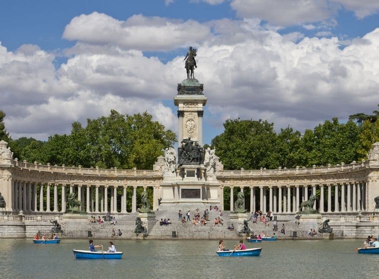 Gent remant amb barques en un llac davant del Monument a Alfons XII al Parc del Retiro de Madrid, amb un teló de fons d'arbres i núvols.