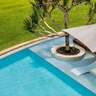 Aerial view of an elegant swimming pool with light blue water at a luxury villa in Mauritius. Two modern white sun loungers with a large parasol providing shade are positioned by the pool edge. A palm tree grows from a circular island within the pool, surrounded by a manicured green lawn creating a tranquil expatriate living environment.