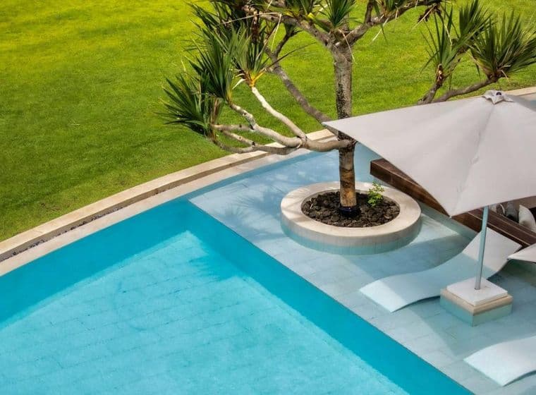 Vue aérienne d'une élégante piscine aux eaux bleu clair dans une villa de luxe à Maurice. Deux transats blancs modernes avec un grand parasol offrant de l'ombre sont positionnés au bord de la piscine. Un palmier pousse depuis un îlot circulaire au milieu de la piscine, entouré d'une pelouse verte bien entretenue, créant un environnement de vie tranquille pour expatriés.