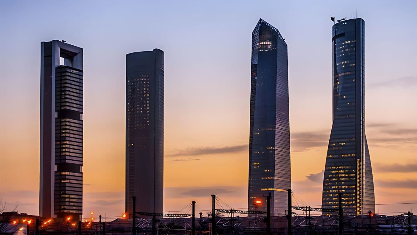 Four modern skyscrapers stand against a sunset sky, with their lights illuminating. The foreground shows structures silhouetted against the skyline.