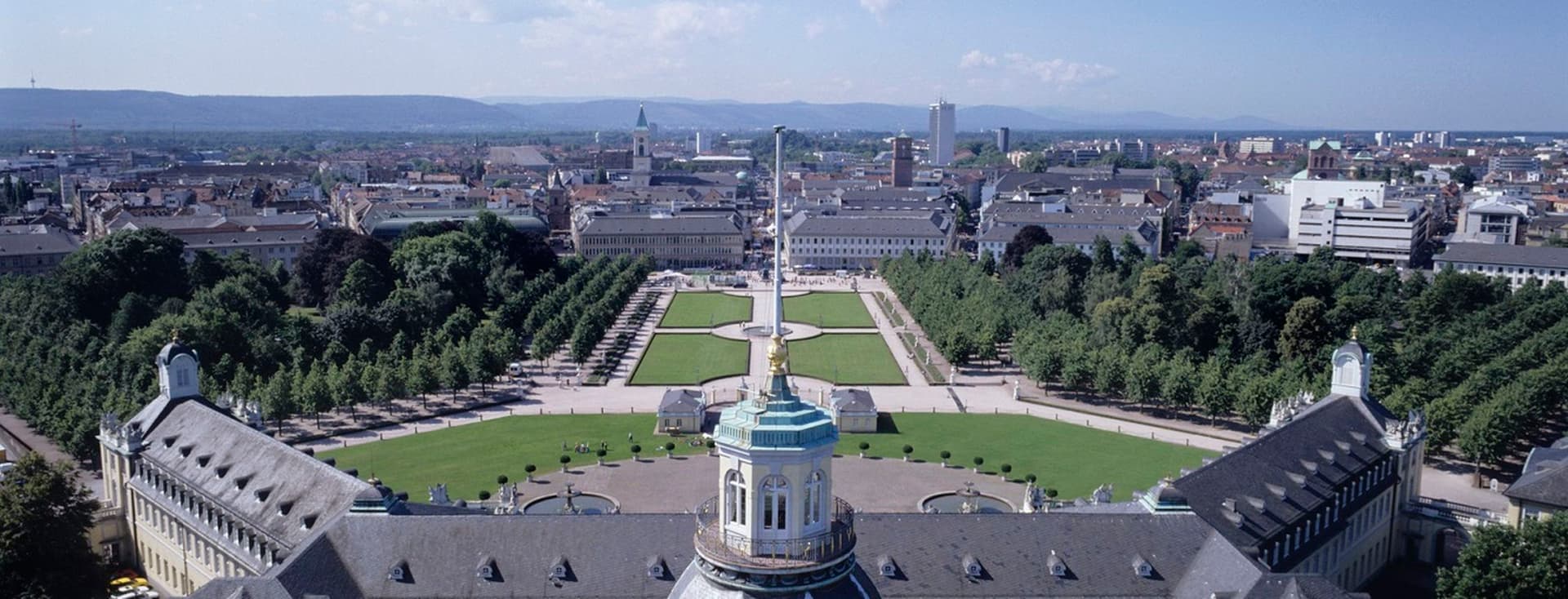 Aerial view of a castle with a large dome in the foreground, surrounded by symmetrically laid-out baroque gardens. Behind the castle stretches a town with numerous buildings, rows of trees, squares, and a hilly landscape in the background under a blue sky with a few clouds.