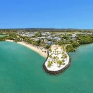 Aerial view of a coastal development in Mauritius showcasing smart urban planning. The image captures a peninsula with pristine white sandy beaches surrounded by turquoise waters. Modern residential buildings are integrated with green spaces and palm trees, demonstrating sustainable coastal development. This bird's-eye perspective illustrates how Mauritius is evolving with smart city initiatives while preserving its natural coastal beauty.