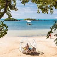 Idyllic beach view in Mauritius framed by tropical trees. White sandy shore with a unique basket-style beach lounger in the foreground. Crystal-clear turquoise waters stretch to the horizon with a small island visible in the distance. A boat floats on the calm sea and a person walks along the shoreline, embodying the peaceful lifestyle awaiting UK expatriates considering relocation to this tropical paradise.