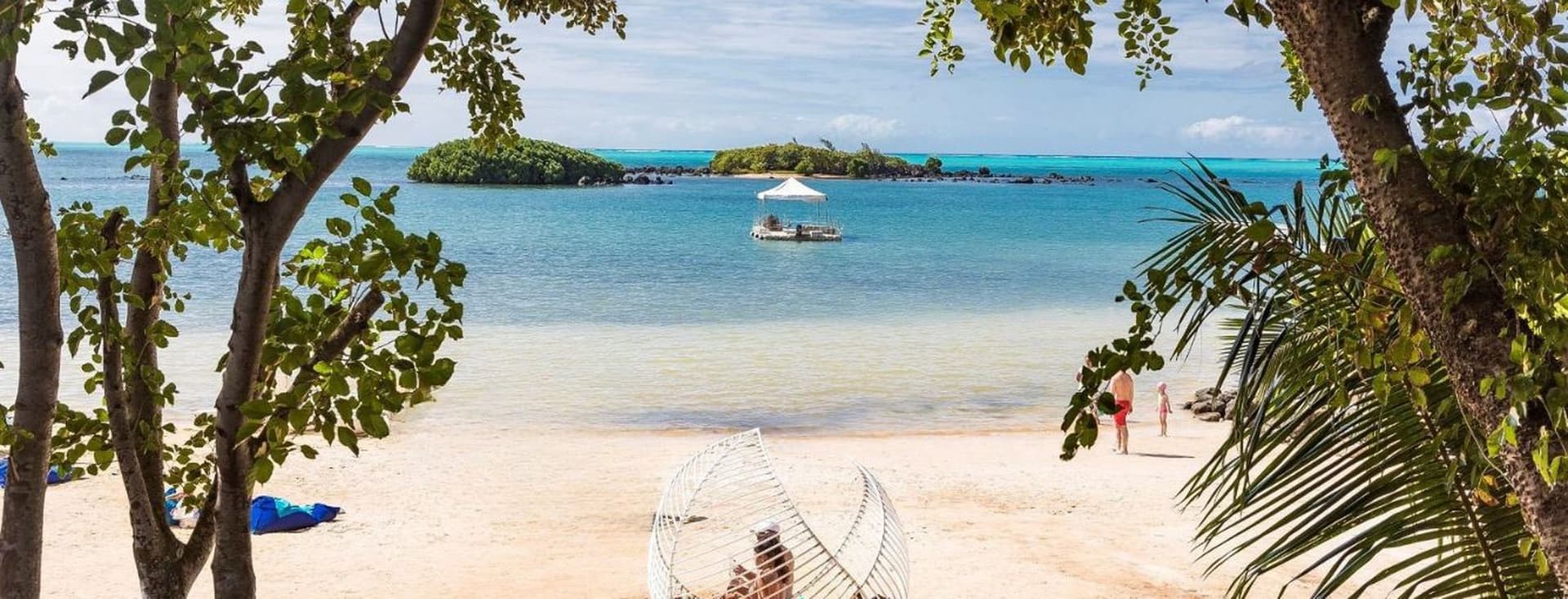 Idyllischer Strandblick auf Mauritius, umrahmt von tropischen Bäumen. Weißer Sandstrand mit einer einzigartigen korbartigen Strandliege im Vordergrund. Kristallklares, türkisfarbenes Wasser erstreckt sich bis zum Horizont mit einer kleinen Insel, die in der Ferne sichtbar ist. Ein Boot schwimmt auf dem ruhigen Meer und eine Person geht am Ufer entlang - verkörpert den friedlichen Lebensstil, der britische Auswanderer erwartet, die einen Umzug in dieses tropische Paradies in Betracht ziehen.