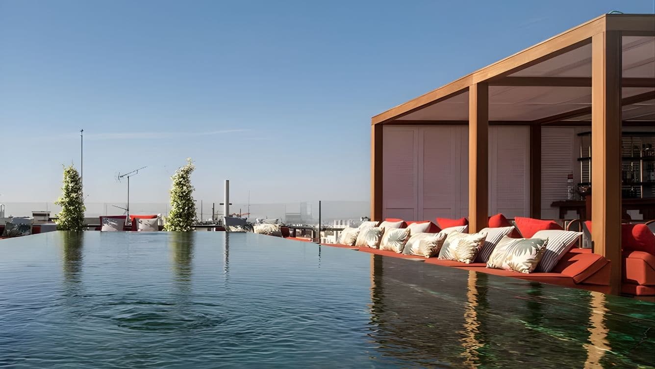 Rooftop infinity pool with lounge area, red cushions, and clear sky in the background. Elegant and serene setting.