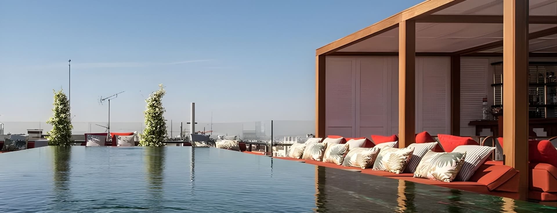 Rooftop infinity pool with lounge area, red cushions, and clear sky in the background. Elegant and serene setting.