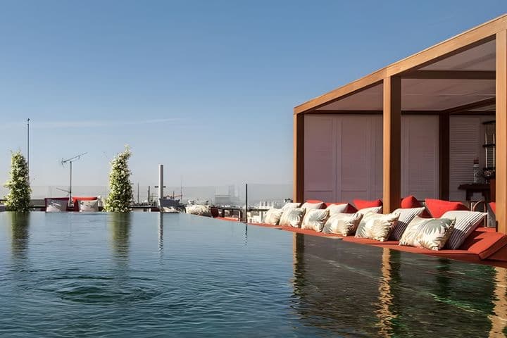 Piscina infinita en la azotea con zona de descanso, cojines rojos y cielo despejado de fondo. Un entorno elegante y sereno.
