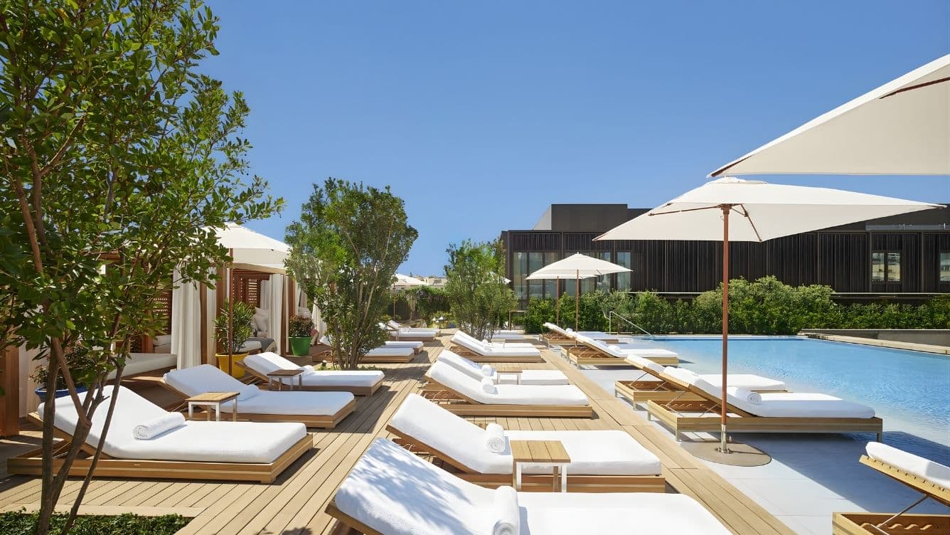 Poolside deck with white loungers, rolled towels, and large umbrellas under a clear blue sky. Trees and greenery surround the area.