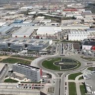 Vista aérea de una zona industrial con fábricas, almacenes, una rotonda y coches aparcados. Paisaje urbano al fondo.