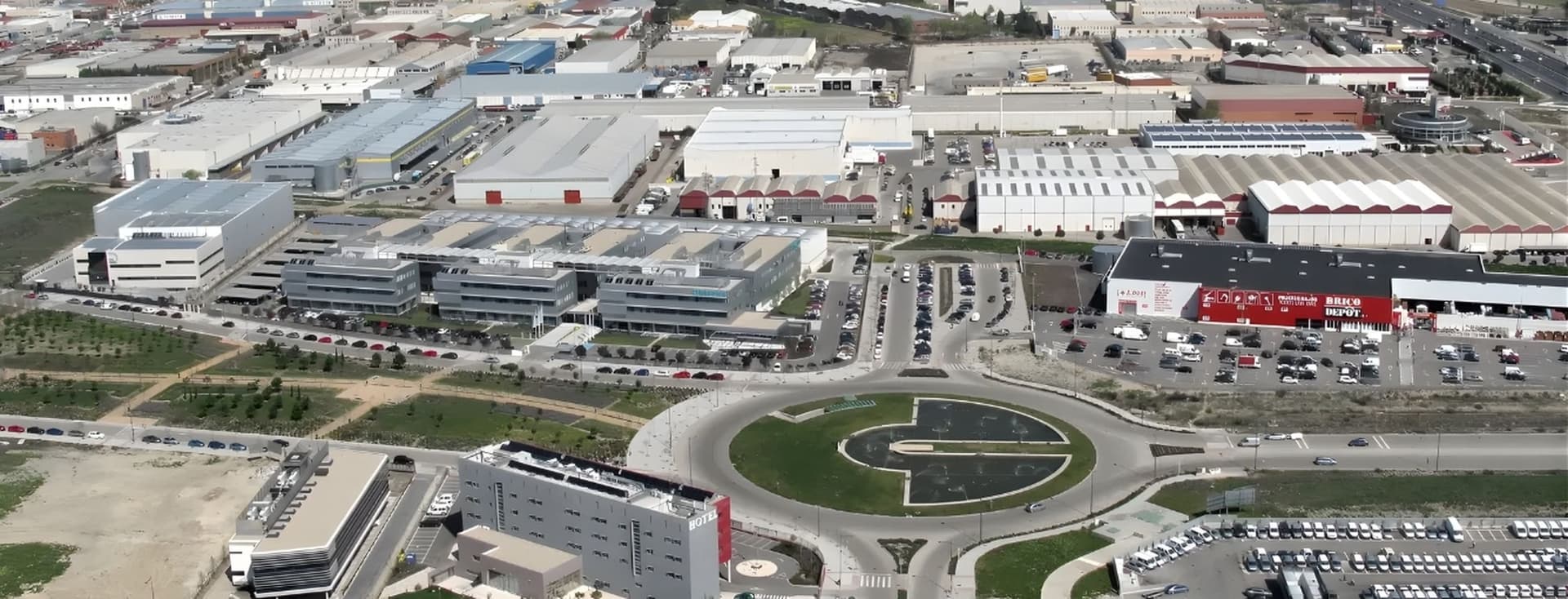 Luftaufnahme eines Industriegebiets mit Fabriken, Lagerhallen, einem Kreisverkehr und geparkten Autos. Im Hintergrund städtische Landschaft.