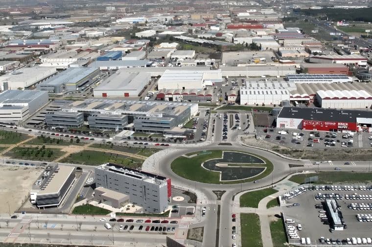 Vista aérea de una zona industrial con fábricas, almacenes, una rotonda y coches aparcados. Paisaje urbano al fondo.
