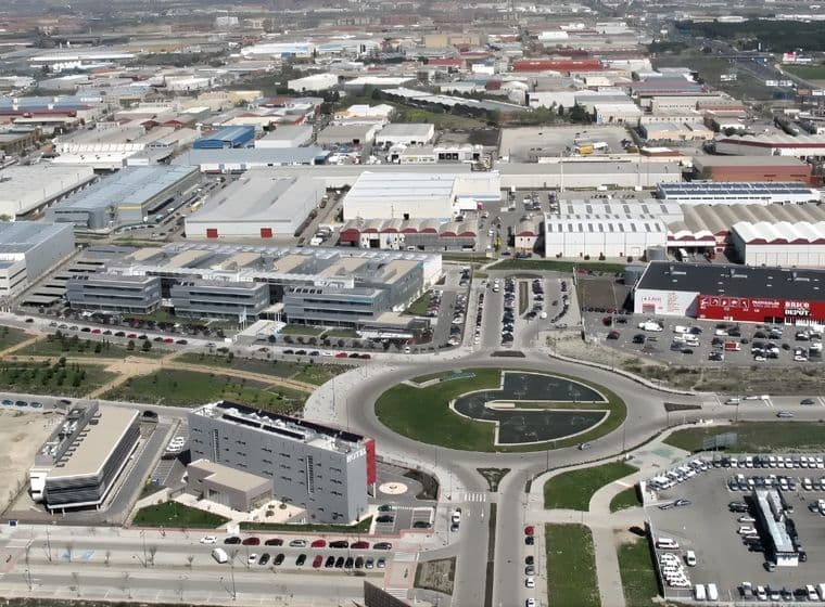 Vista aérea de una zona industrial con fábricas, almacenes, una rotonda y coches aparcados. Paisaje urbano al fondo.