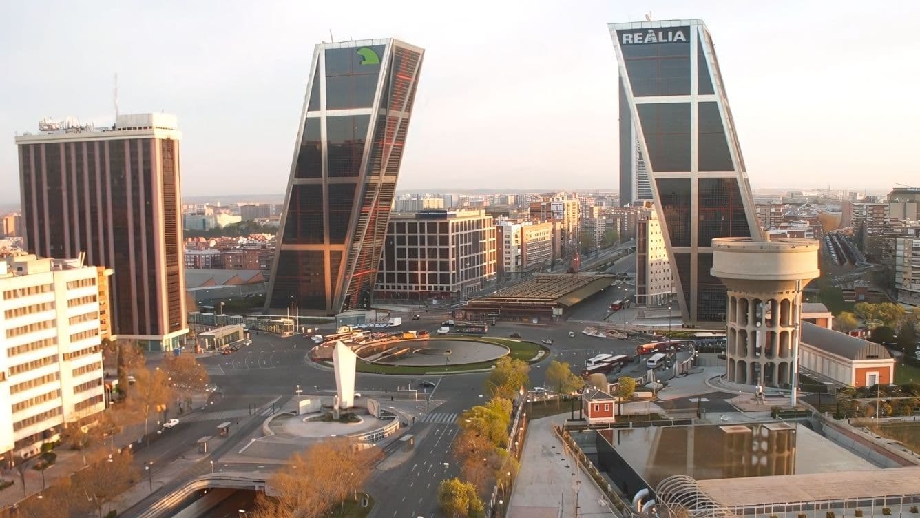 Cityscape featuring the leaning KIO Towers in Madrid, surrounded by urban buildings, roads, and a roundabout with a monument.