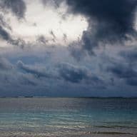 Dramatic stormy sky over the coast of Mauritius during cyclone season. Dark, turbulent clouds dominate the image, creating an ominous atmosphere above the otherwise calm turquoise sea. The contrast between the threatening sky and peaceful ocean highlights the unpredictable nature of weather during cyclone season, serving as a visual reminder for expatriates about the importance of storm preparedness and safety measures.