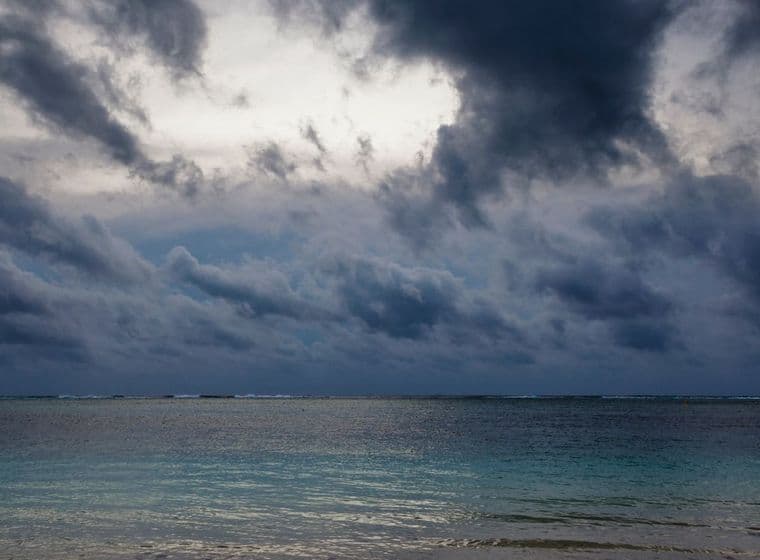 Dramatischer, stürmischer Himmel über der Küste von Mauritius während der Zyklonsaison. Dunkle, turbulente Wolken dominieren das Bild und schaffen eine bedrohliche Atmosphäre über dem ansonsten ruhigen, türkisfarbenen Meer. Der Kontrast zwischen dem bedrohlichen Himmel und dem friedlichen Ozean unterstreicht die Unberechenbarkeit des Wetters während der Zyklonsaison und dient als visuelle Erinnerung für Expatriates an die Wichtigkeit von Sturmvorbereitungen und Sicherheitsmaßnahmen.