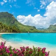 Breathtaking coastal landscape of Mauritius featuring a pristine beach with crystal-clear turquoise waters. The scene is framed by vibrant pink bougainvillea flowers in the foreground, lush palm trees along the shoreline, and dramatic green mountains in the background. Fluffy white clouds dot the bright blue sky above the verdant peaks, showcasing the natural beauty that makes this island an idyllic place to live.