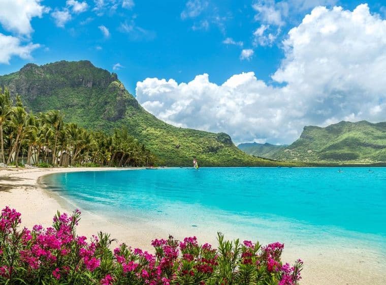 Breathtaking coastal landscape of Mauritius featuring a pristine beach with crystal-clear turquoise waters. The scene is framed by vibrant pink bougainvillea flowers in the foreground, lush palm trees along the shoreline, and dramatic green mountains in the background. Fluffy white clouds dot the bright blue sky above the verdant peaks, showcasing the natural beauty that makes this island an idyllic place to live.