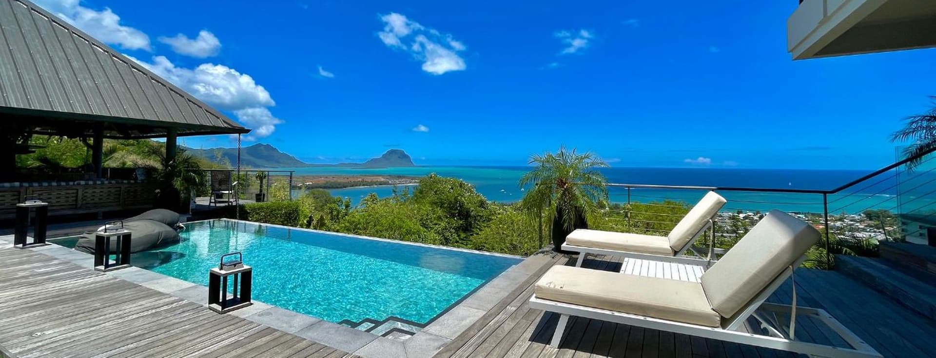Une élégante villa de luxe à l'Île Maurice dotée d'une splendide piscine à débordement surplombant l'océan turquoise. La terrasse en bois entoure la piscine avec des chaises longues confortables, tandis que la végétation tropicale encadre la vue sur une petite île visible au loin sous un ciel bleu éclatant parsemé de nuages.