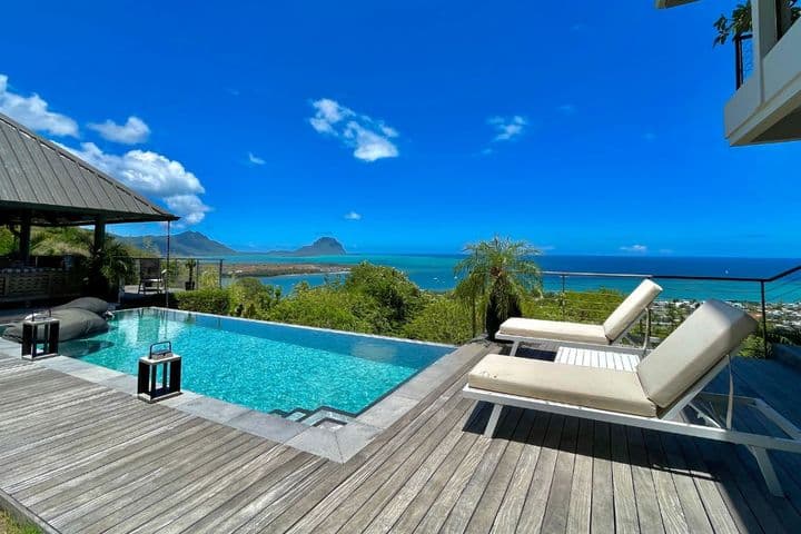 Eine elegante Luxusvilla auf Mauritius mit einem atemberaubenden Infinity-Pool mit Blick auf den türkisfarbenen Ozean. Die Holzterrasse umgibt den Pool mit komfortablen Liegestühlen, während tropische Vegetation den Ausblick auf eine kleine, in der Ferne sichtbare Insel unter einem strahlend blauen Himmel mit vereinzelten Wolken einrahmt.