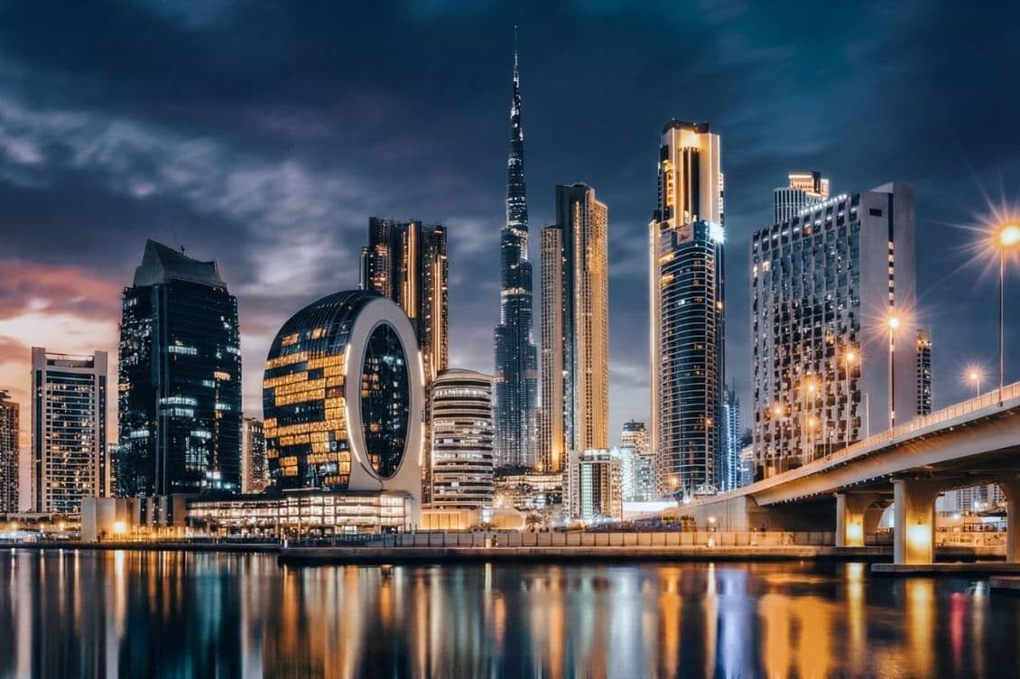 Dubai skyline at night with illuminated skyscrapers, including a distinctive circular building, reflected in calm water under a cloudy sky.