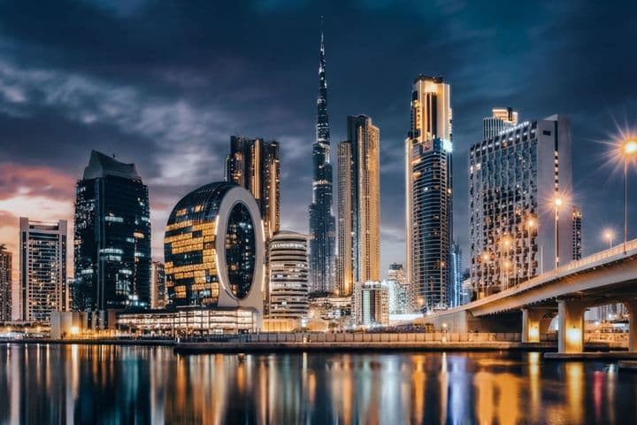 Dubai skyline at night with illuminated skyscrapers, including a distinctive circular building, reflected in calm water under a cloudy sky.