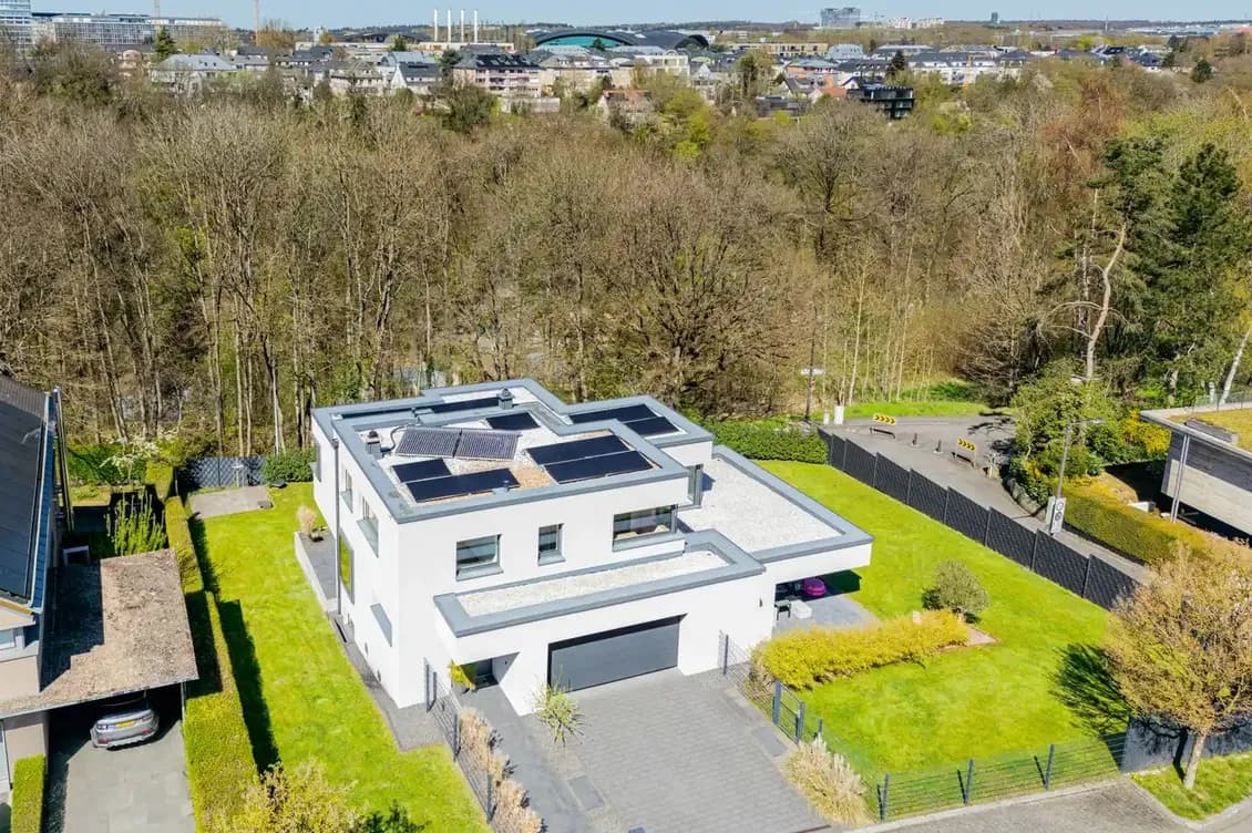 Modern white two-story house with a flat roof and solar panels, a gray driveway to a double garage, and a green lawn beside trees.